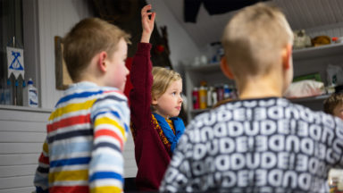 Kaksi pientä lasta selkä kameraan päin värikkäät paida päällä. Huivikaulainen tyttö viittaa taustalla