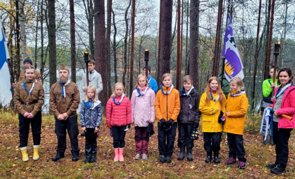 Aikuisia ja lapsia rivissä partiohuivit kaulalla Suomenlipun ja Metsänkkävijöiden lipun sekä palavien soihtujen ollessa taustalla.