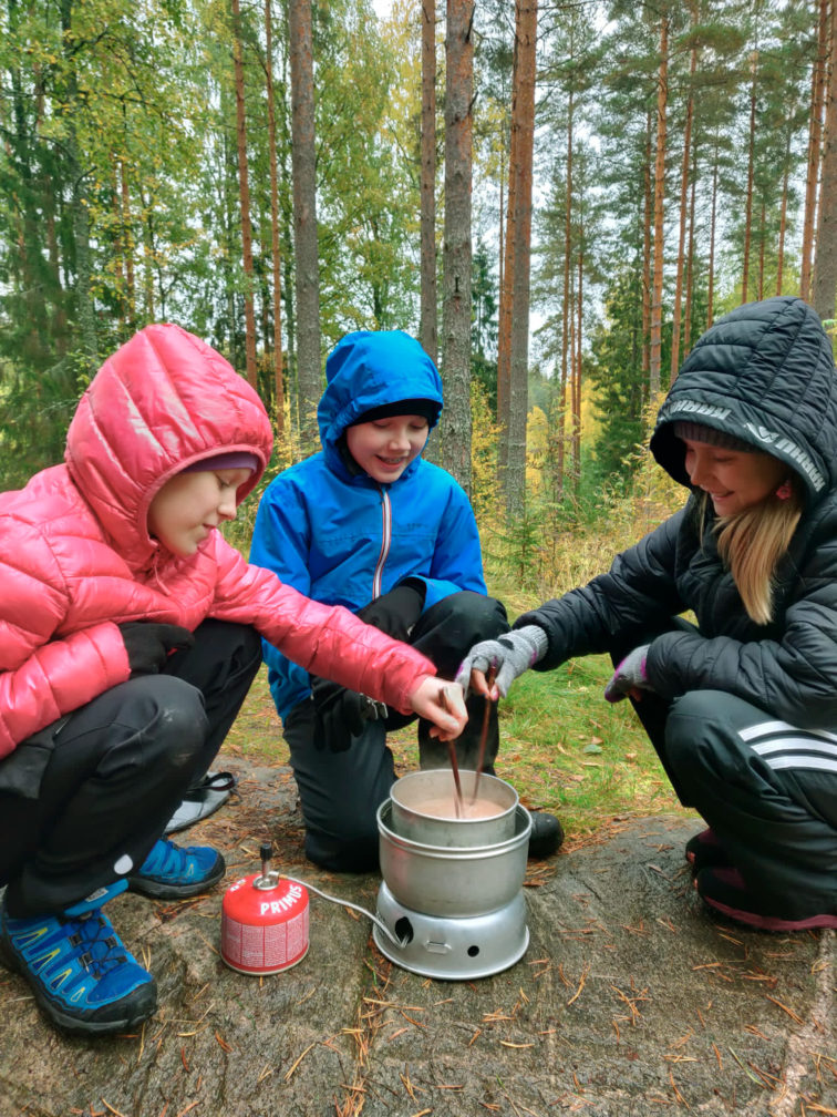 Kolme tyttöä metsässä syyssäässä keittämässä kaakaota retkikeittimellä.