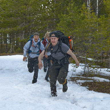 Kolme nuorta miestä juoksemassa rinkat selässä lumisessa metsässä partiotaitokisojen seuraavalle rastipaikalle.