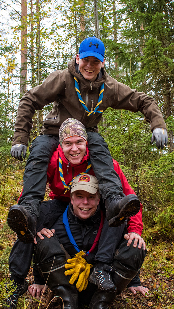 Kolme nuorta miestä istuvat toistensa olkapäillä metsässä ja nauravat.