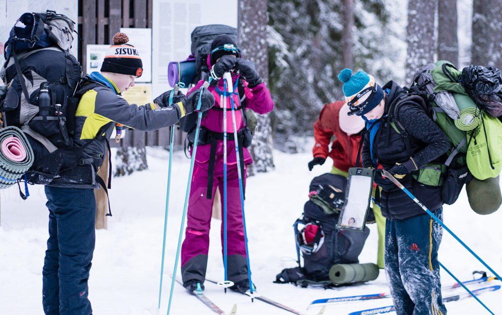 Kolme partiolaista syksien päällä laittamassa varusteita tiukalle hiihtämään lähdössä. Talvisessa maisemassa neljäs pakkaa taustalla vielä rinkkaa.
