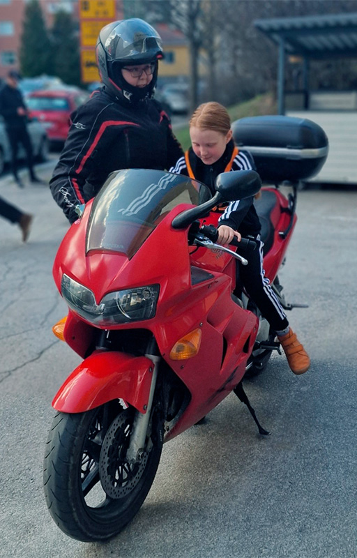 Kypäräpäinen naismotoristi tukee sivusta punaista moottoripyörää, jonka päällä istuu punatukkainen, mustaan verryttelyasuun pukeutunut seikkailijatyttö.